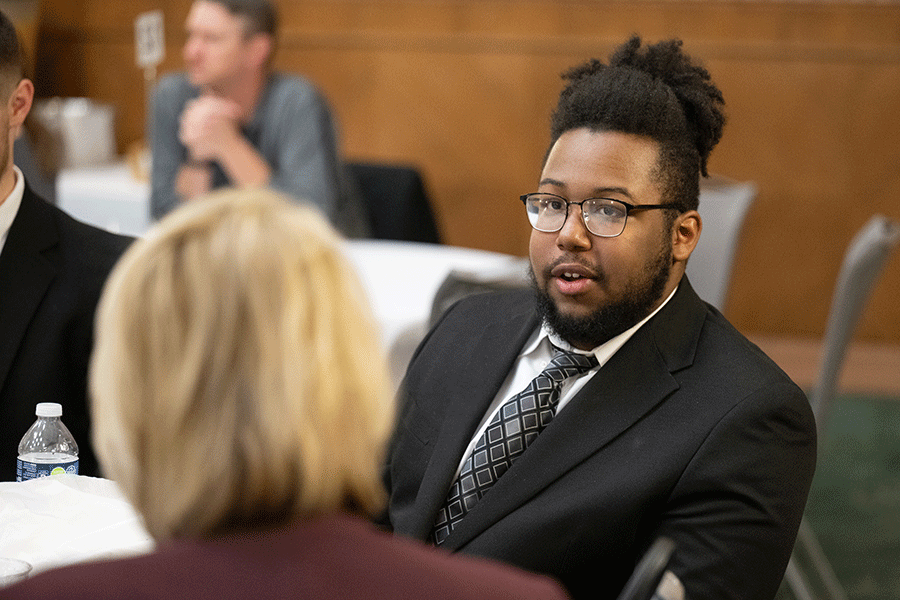 A man in a black business suit with white shirt and grey checkered tie is seated at a table in a conference room. He is talking with another person whose face is not visible.