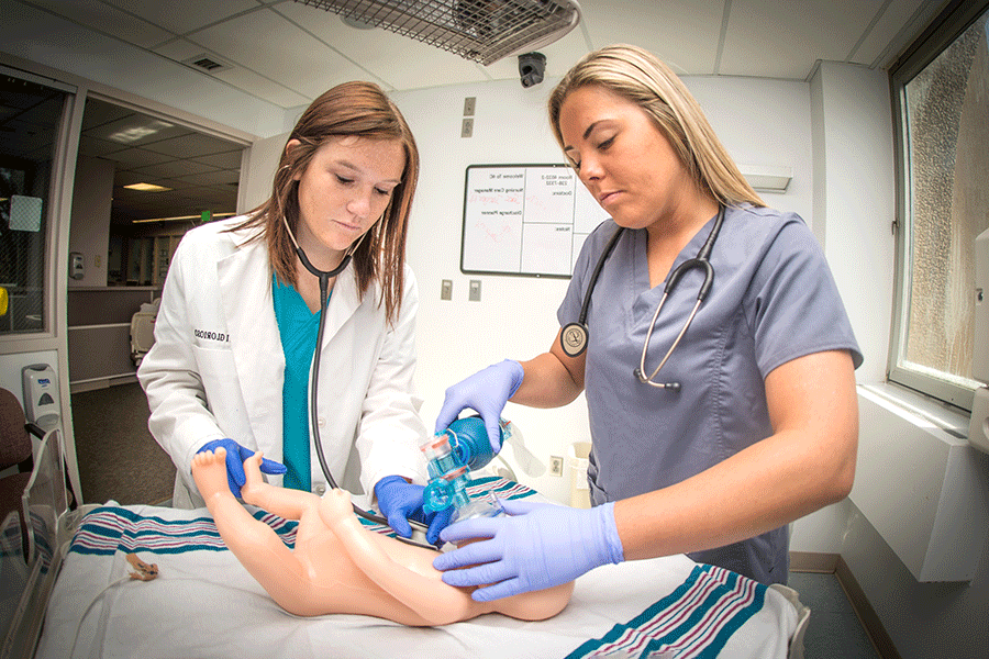 Two female students in a medical room. They are looking at an infant mannequin. On the right, a female student with brown hair wears teal scrubs, a white lab coat, and a stethoscope. On the left, a female student with blonde hair wears blue scrubs. She holds an oxygen mask to the mannequin.