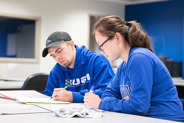 Two students, an woman with dark hair in a ponytail and glasses, and a man wearing a grey baseball cap, work on an assignment together. They both wear blue ISU apparel, and both hold writing implements in their hands.