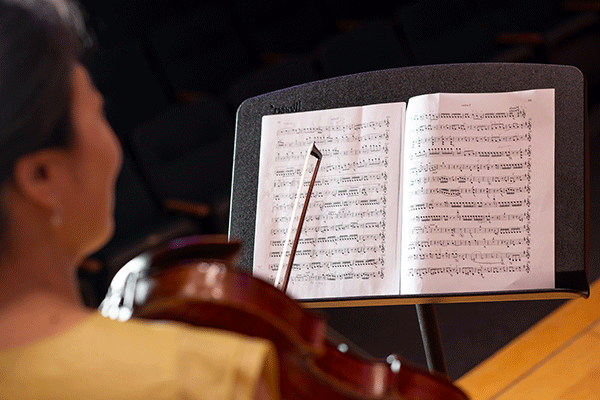 A book of sheet music is open on a black music stand. Blurred in the foreground, a woman with short black hair wearing a white blouse is holding a violin on her shoulder, with the bow extending up from her other hand. The background of the image is very dark.