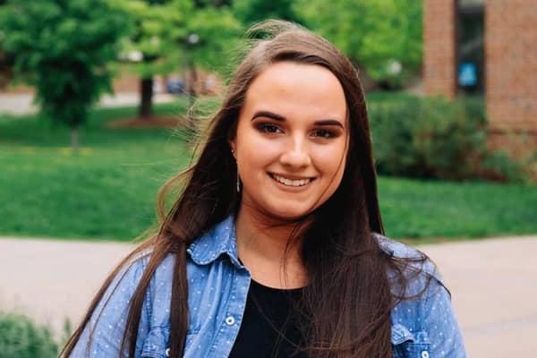 A white female student with long brown hair poses outside. She wears a blue jean jacket with a black undershirt. A brick building, a sidewalk, and a lawn with green trees are blurred in the background.