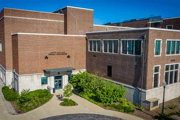 aerial image of a red-and-white brick, multi-story building that houses the Norma and William Grosjean Clinic. The clinc’s name is visible above the door. There is blue sky visible above and green grass and trees visible below. 