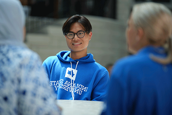 A male student of Asian descent with short dark hair, glasses, and a blue hoodie with Indiana State University in white lettering smiles while sitting at a table. He talks to two female students with their backs to the camera.  