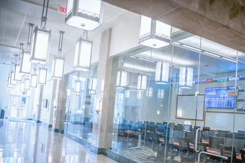 brightly lit hallway of a building with a a classroom/lab on the left side of the frame with glass doors and windows allowing the viewer to see a trading ticker and large screens I the room.