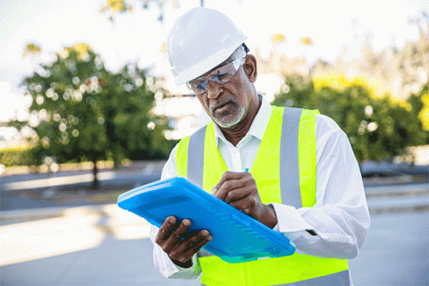 A senior black male construction site manager writing on a blue tablet while wearing a hard hat, safety goggles and a yellow construction vest while inspecting a building project